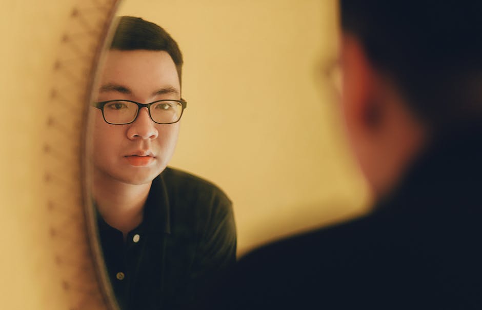 East Asian man with glasses looking at his reflection in a mirror, contemplative mood.